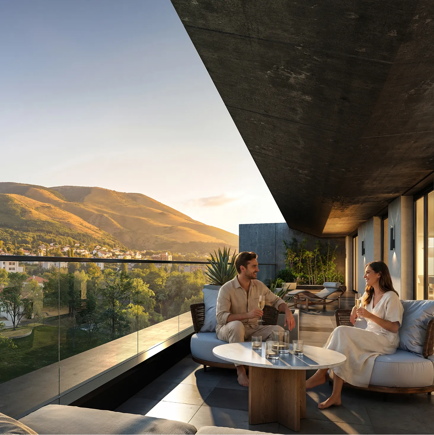 A man and woman sit on a modern balcony with drinks, overlooking a scenic landscape with hills and trees at sunset.